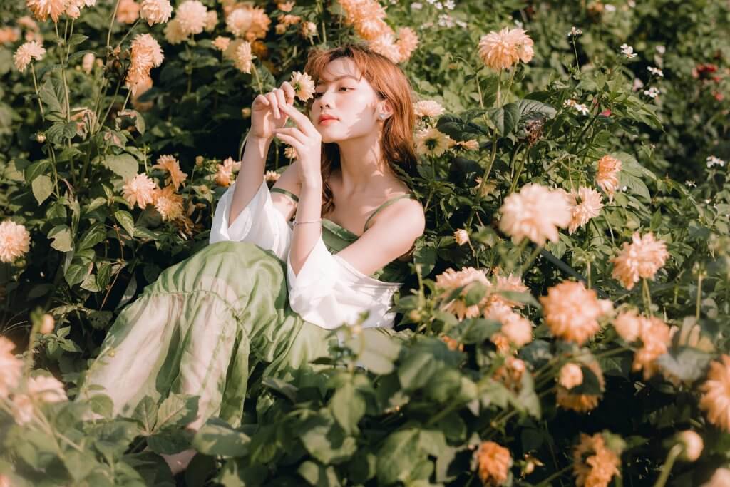 Woman in white dress sits in green field with light pink flowers