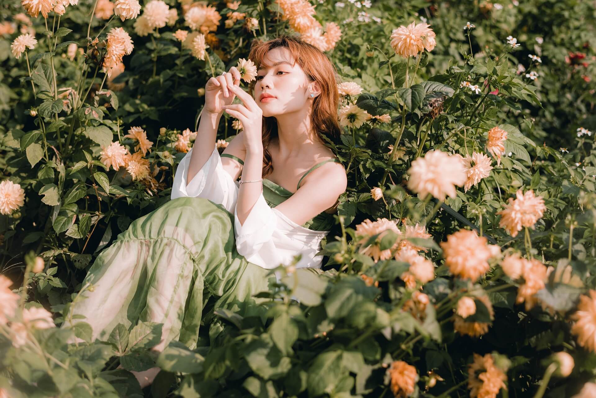 Woman in white dress sits in green field with light pink flowers