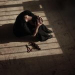 Woman sits on the floor inside with sun shining through the window on her and holding and hiding her head