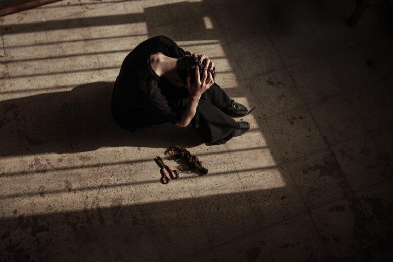 Woman sits on the floor inside with sun shining through the window on her and holding and hiding her head